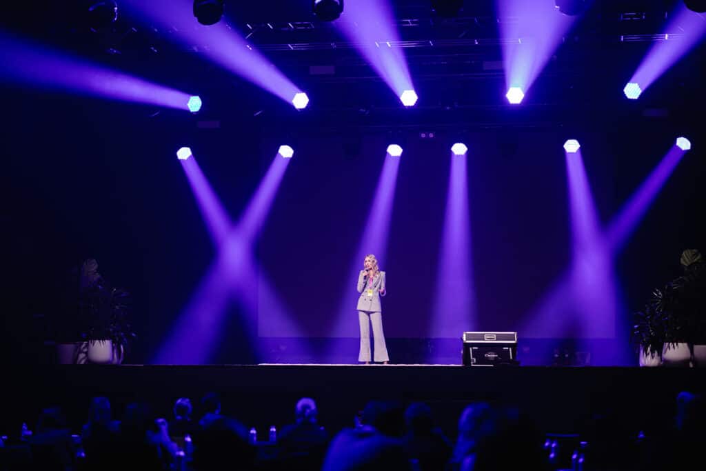 An Image of WDEN Founder Anja Christoffersen on stage, with the audience blurred in the foreground. The stage has bright blue lights, and Anja stands in the centre with a microphone speaking, wearing a black and white patterned suit. Anja has medium-length blonde hair, and is holding the microphone with one hand, extending her other hand to the audience.